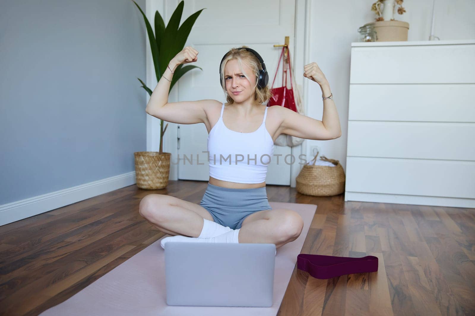 Portrait of young sporty woman following online video instructions during fitness workout, using laptop, sitting on yoga mat by Benzoix