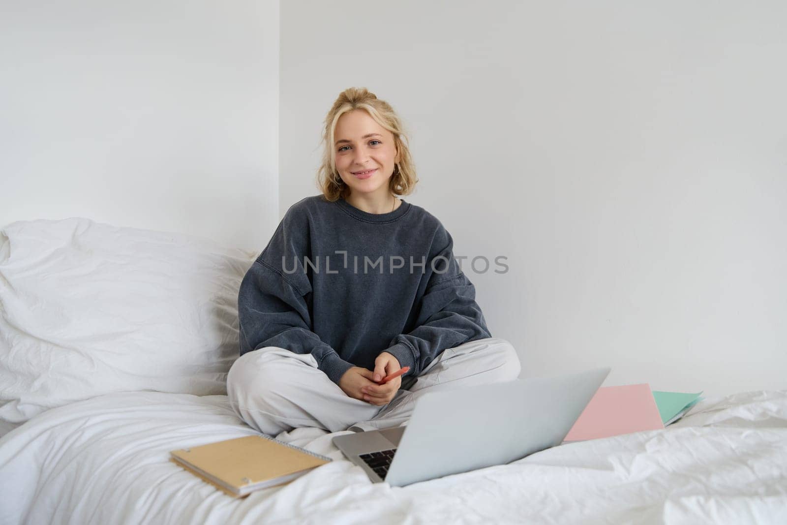 Portrait of female student, woman sits on bed with laptop and notebooks, studying online, remote education concept. Girl freelancer working from her bedroom.