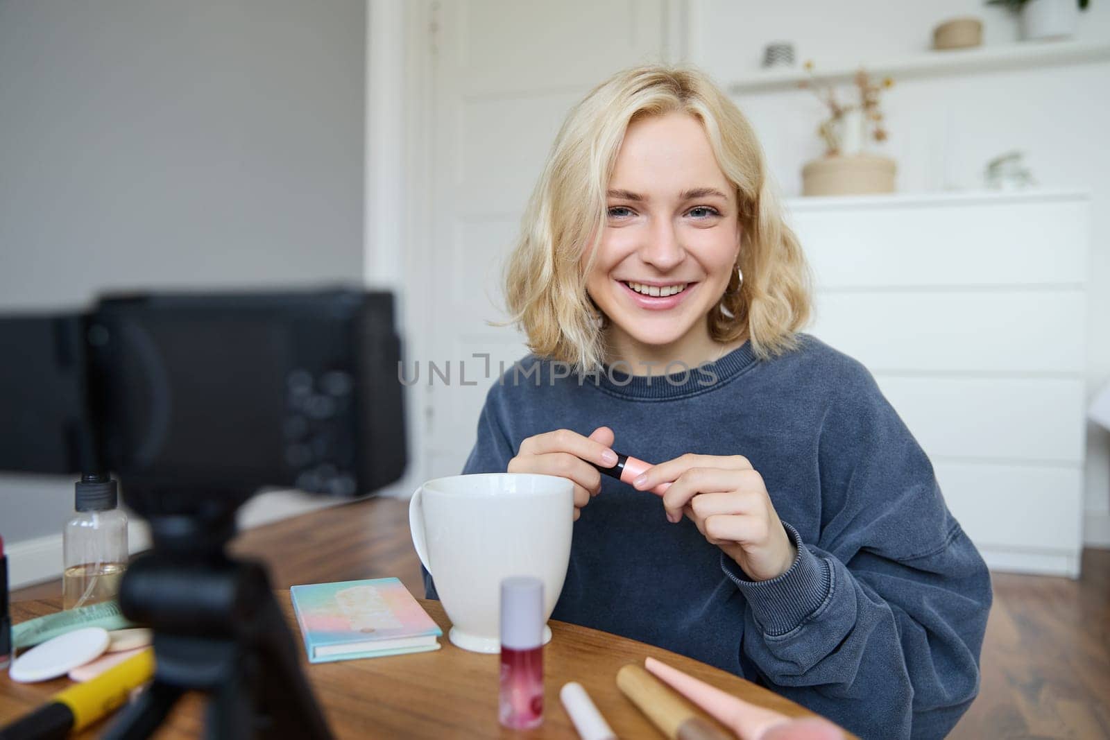 Portrait of beautiful, smiling blond woman, girl recording video of her makeup tutorial for social media, vlogger sitting on floor in her room, using stabiliser to create content, reviewing mascara.