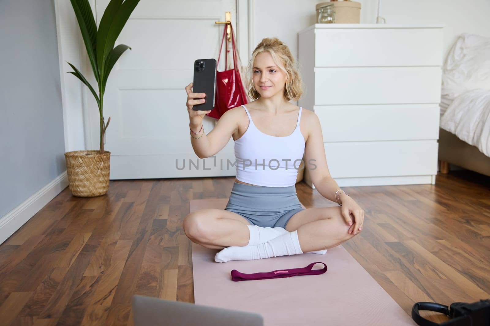 Portrait of stylish young woman, sitting on rubber mat on floor at home, taking selfie while doing workout, yoga session, posting photo on social media by Benzoix