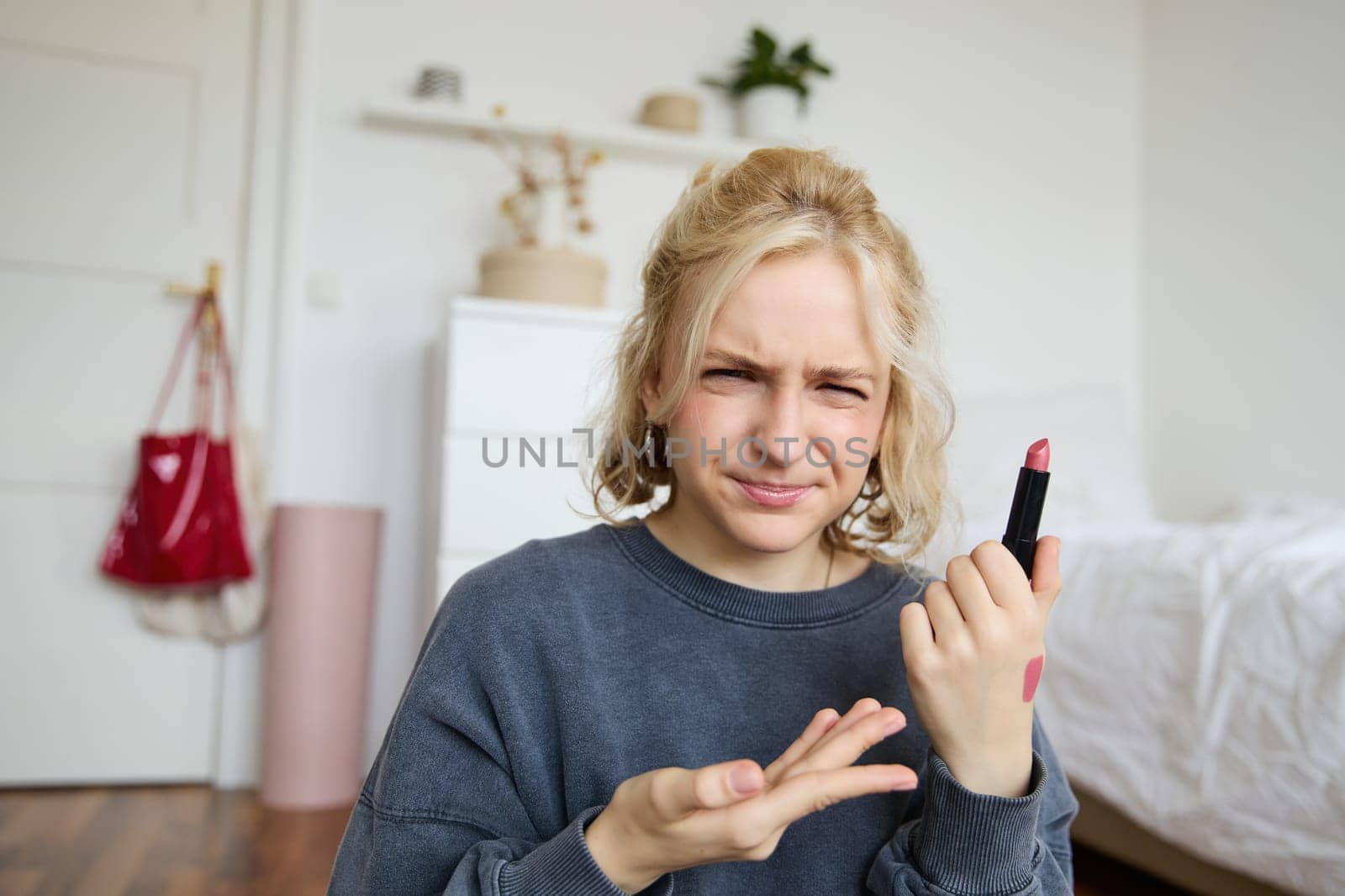 Portrait of young blogger, woman recording video about beauty, makeup products, showing lipstick and grimacing, express negative opinion, creating a lifestyle vlog content.