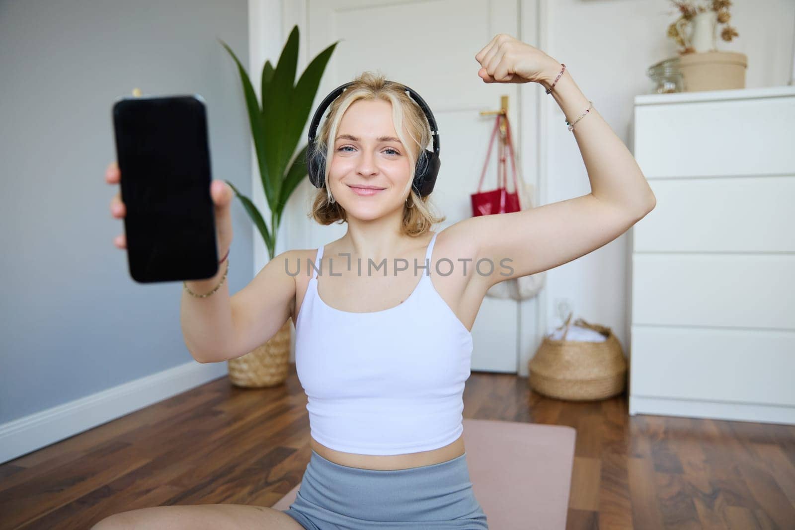 Smiling blond woman working out, showing biceps, recommending smartphone app, demonstrating mobile phone screen at camera, sitting on rubber yoga mat in a room by Benzoix