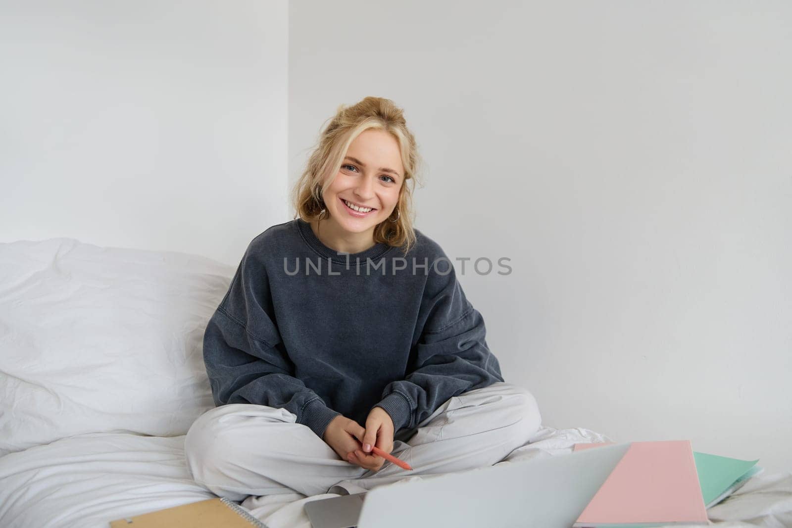 Portrait of young beautiful woman sitting on bed with laptop and notebooks, working from home, freelancing. Female student studying online, video chats, connects to lesson remotely.