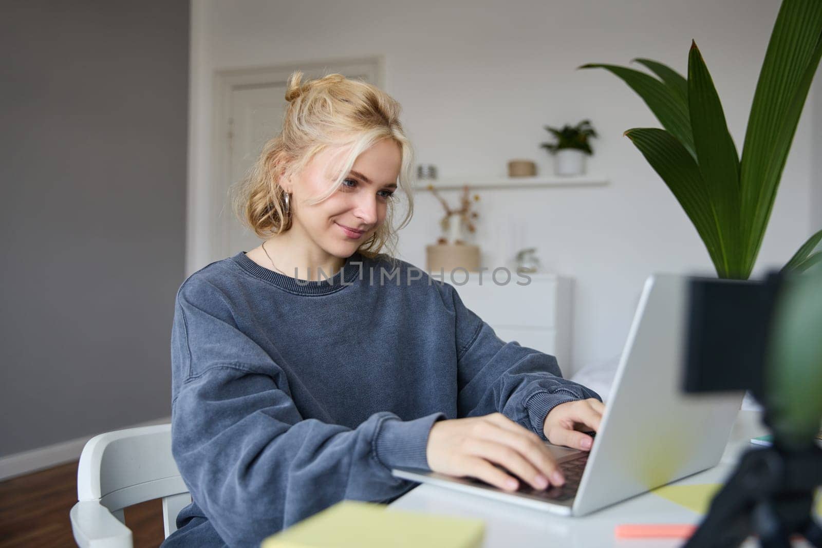Portrait of young blond woman, female college student works from home on assignment, uses laptop, studies remotely, sits in room in front of computer.