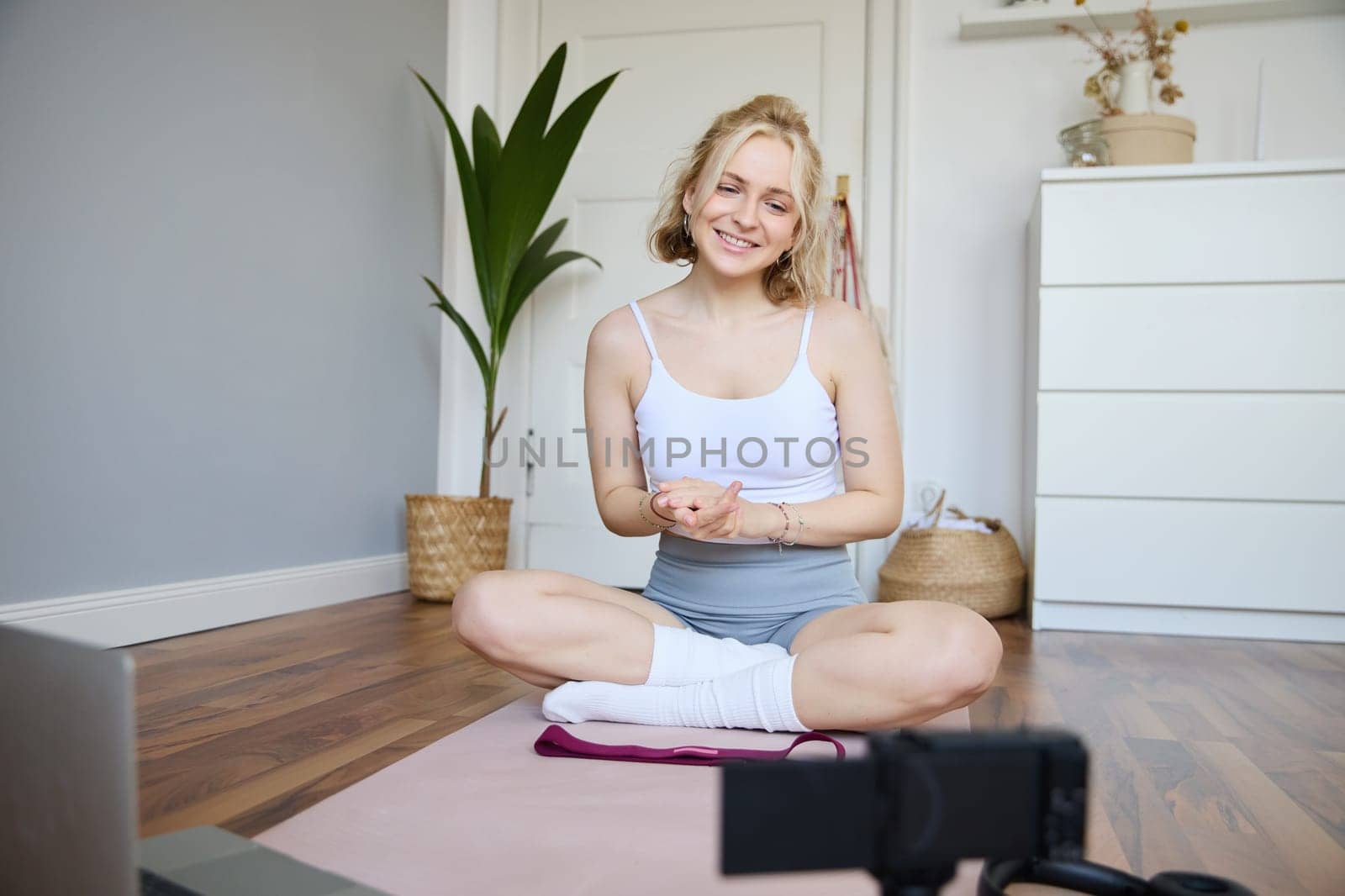 Young female athlete, fitness instructor woman sits on floor rubber mat, recording video on digital camera, showing how to workout, explaining exercises by Benzoix