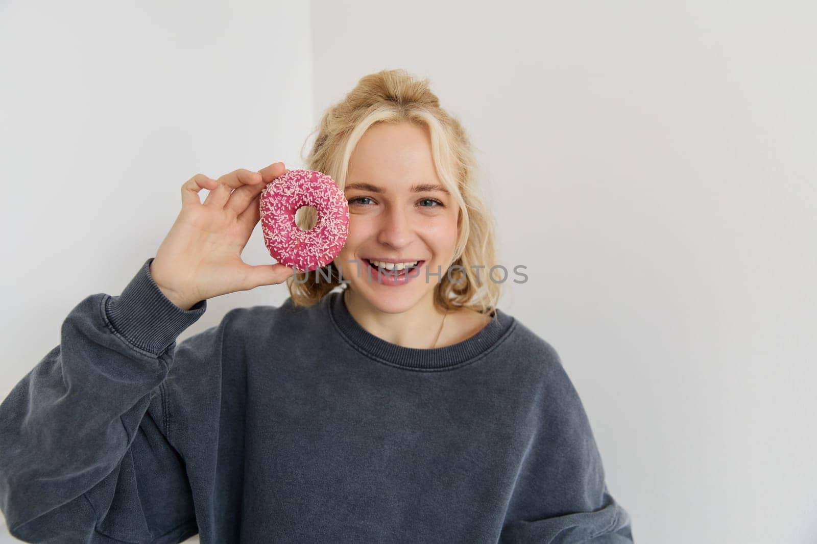 Close up portrait of cute young blond woman, showing pink doughnut near her face, smiling and looking happy by Benzoix