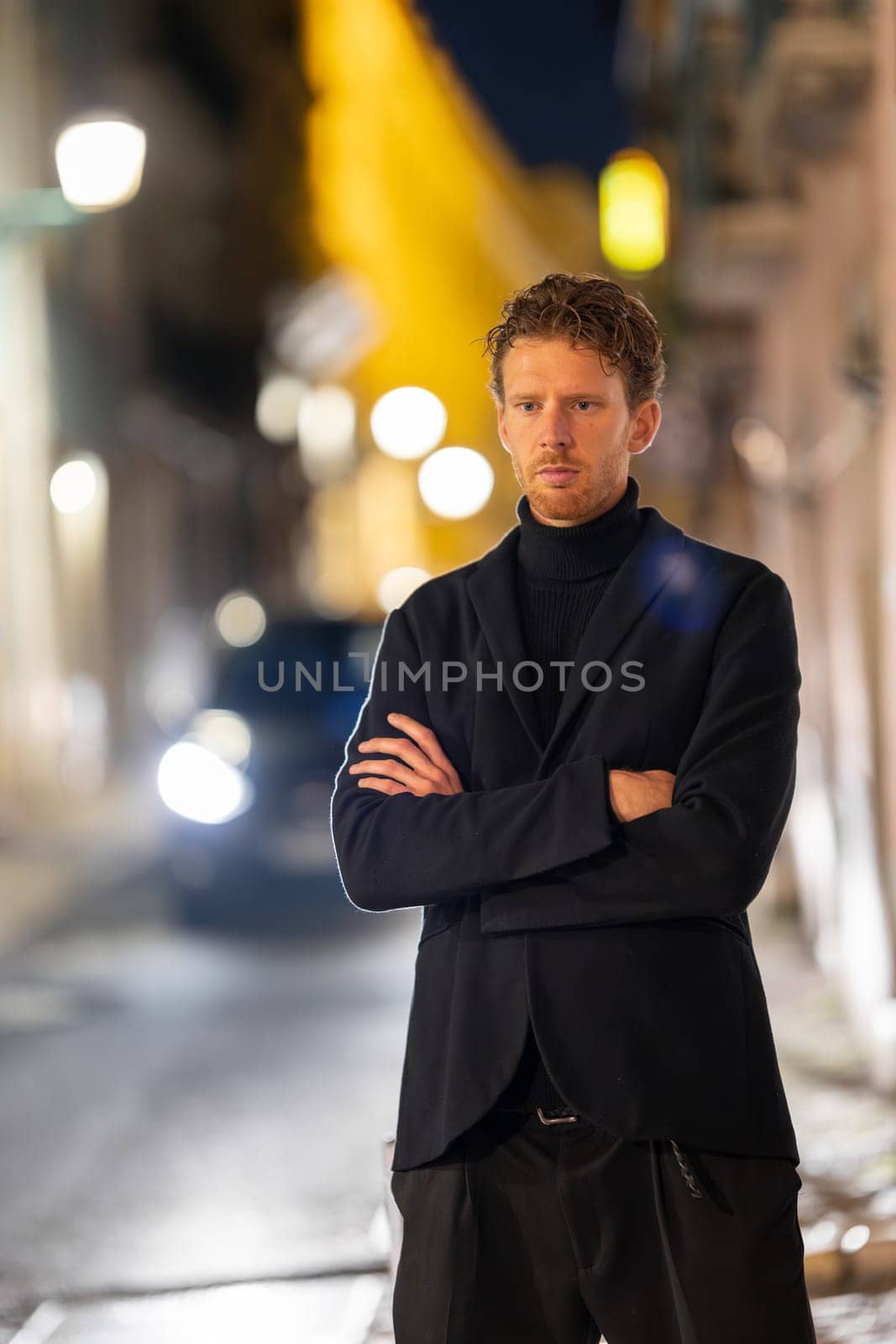 A handsome man is standing in the street with his arms crossed. He is wearing a black jacket and a black tie. The street is lit up with bright lights, creating a moody atmosphere