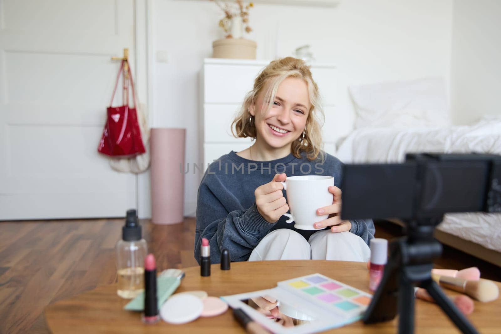 Portrait of smiling young woman, girl records video on camera, holds cup of tea, talking, doing lifestyle blog, sitting in room and creating content.