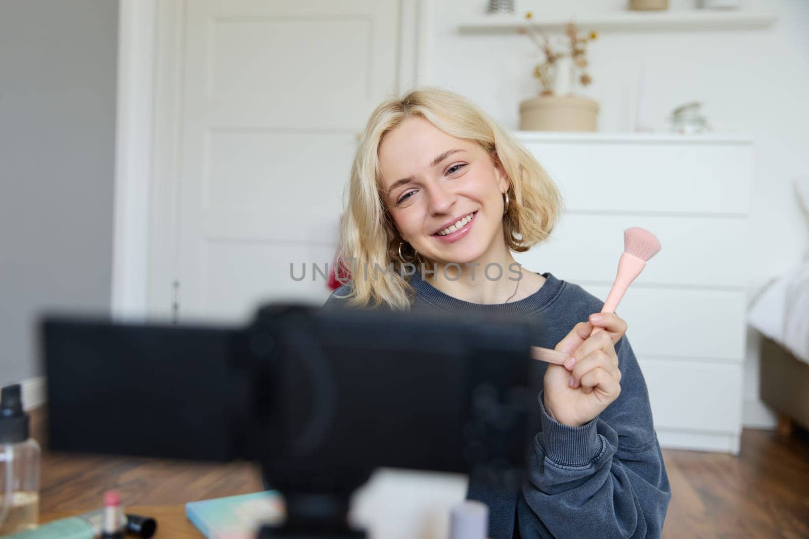 Close up portrait of happy young beauty blogger, records lifestyle vlog in her room, using camera with stabiliser, shows makeup brush and cosmetics.