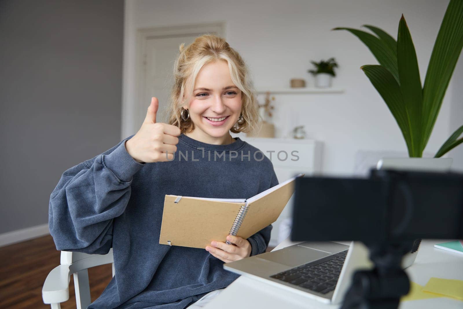 Portrait of smiling blond woman, social media vlogger, reads notes from notebook and looks at digital camera, records video in her room, shows thumbs up.
