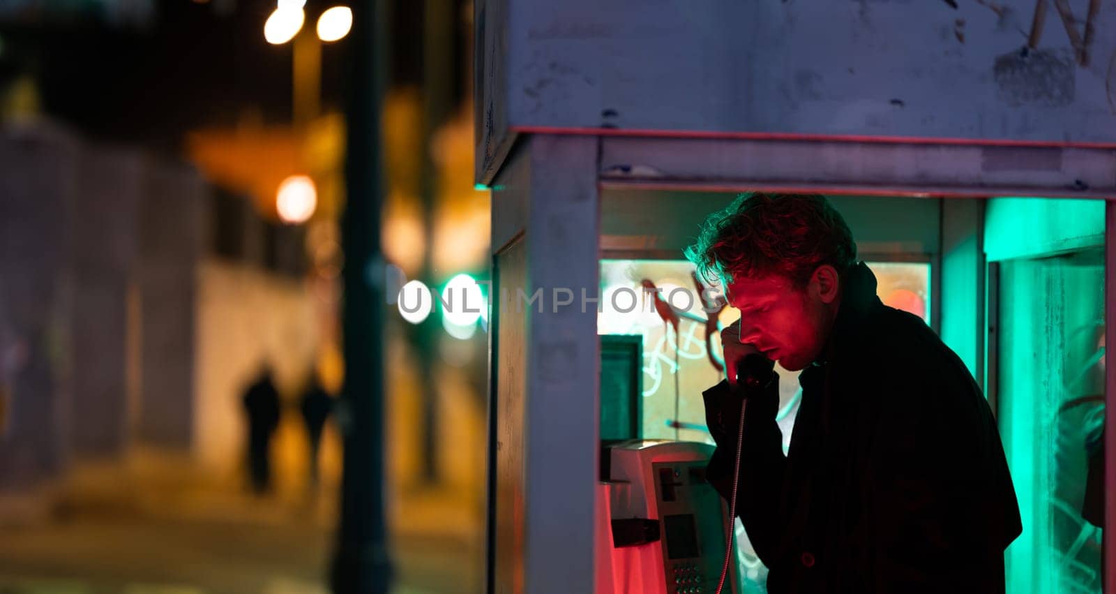 A man is talking on a phone at noir style in booth The image has a dark and moody atmosphere, with the man's face illuminated by the phone's light