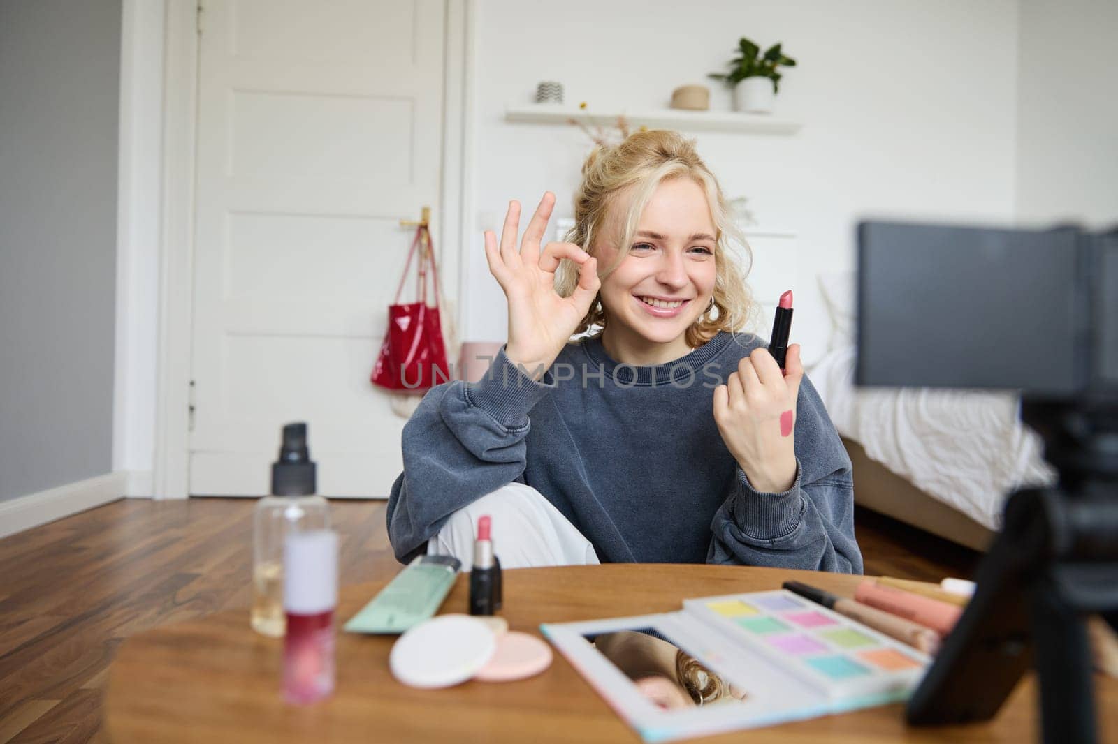 Cheerful, smiling young woman recording a vlog, showing okay, ok gesture and favorite lipstick, recommending beauty product for social media followers, using digital camera to create content.
