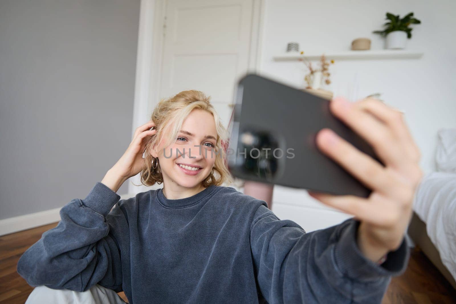 Portrait of young woman, social media influencer, taking selfies in her room, sitting on floor, holding smartphone and posing for a photo.