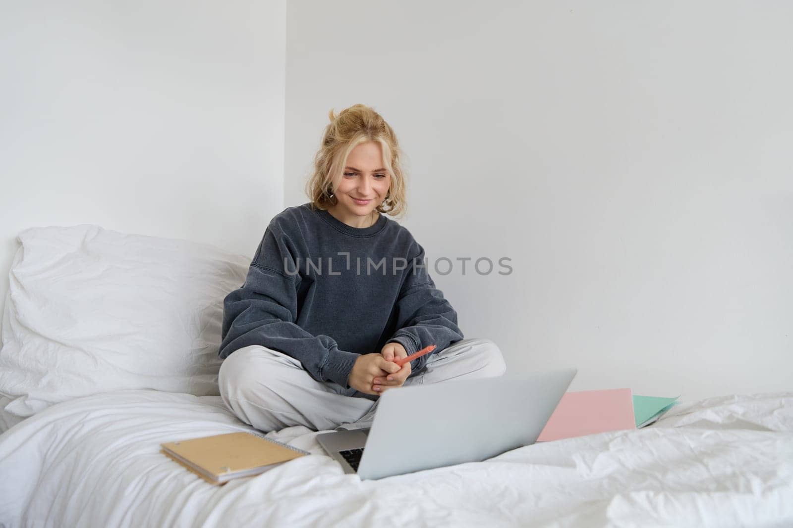 Portrait of happy blond woman, freelancer working from home, sitting on bed with laptop and notebooks. Student doing homework in bedroom, connects to online class via video chat.