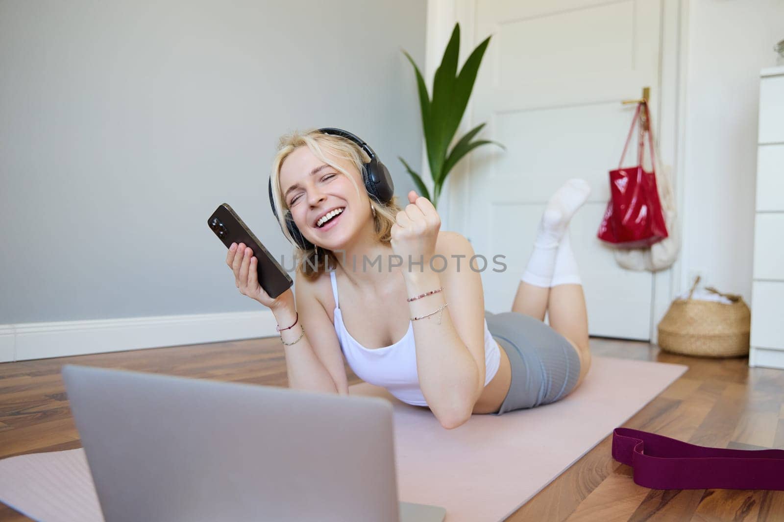 Young woman doing fitness workout at home, lying on rubber yoga mat, holding smartphone, wearing headphones, using laptop to watch sports videos by Benzoix