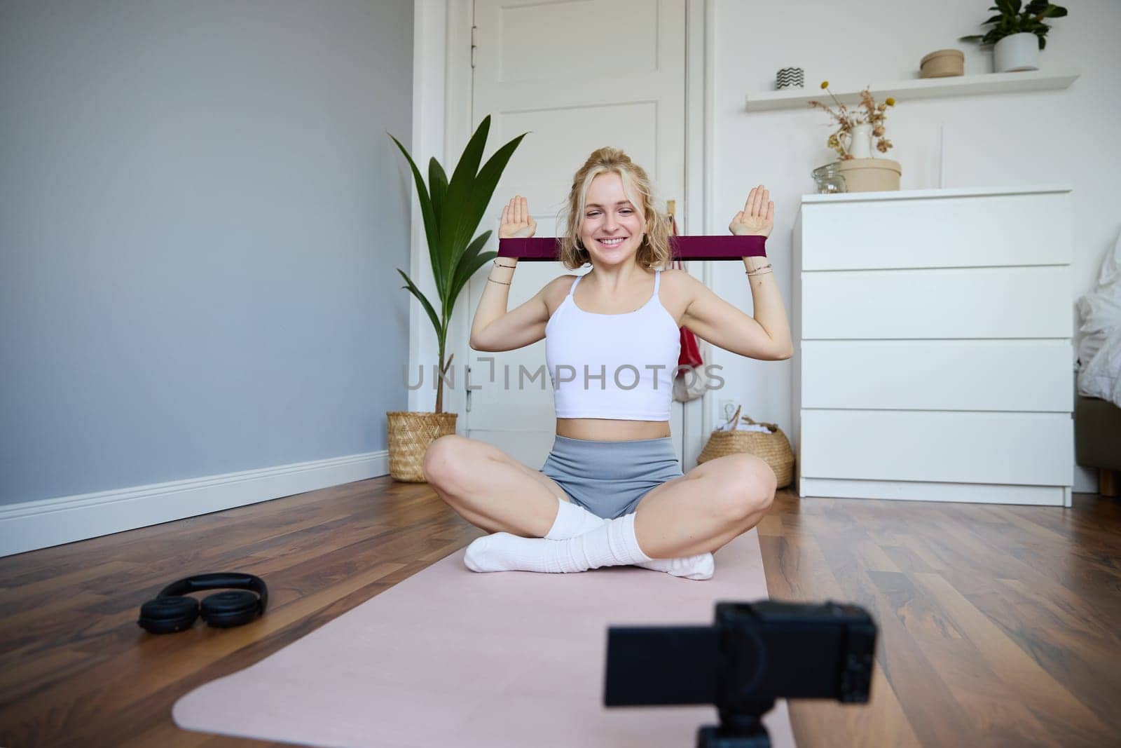 Portrait of young athletic woman recording home workout video, shooting content for sport fitness vlog, using resistance band and digital camera.