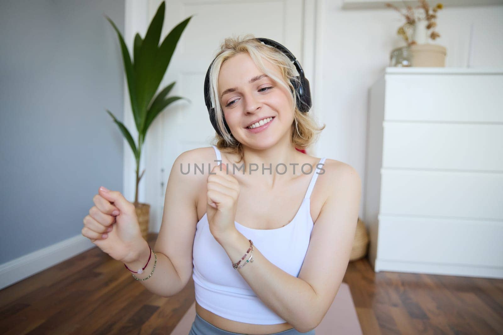 Portrait of happy, beautiful athletic woman, working out at home, listening music to boost energy while active training session, using wireless headphones and rubber yoga mat.