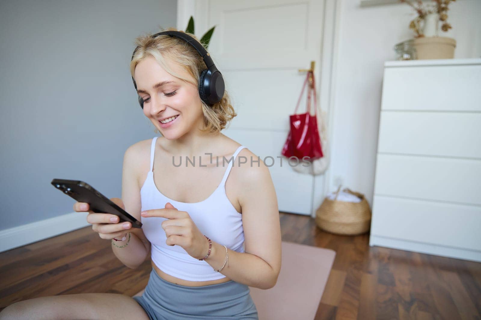 Close up portrait of blond young woman choosing fitness podcast, looking at her mobile phone, checking workout app on smartphone, wearing wireless headphones.