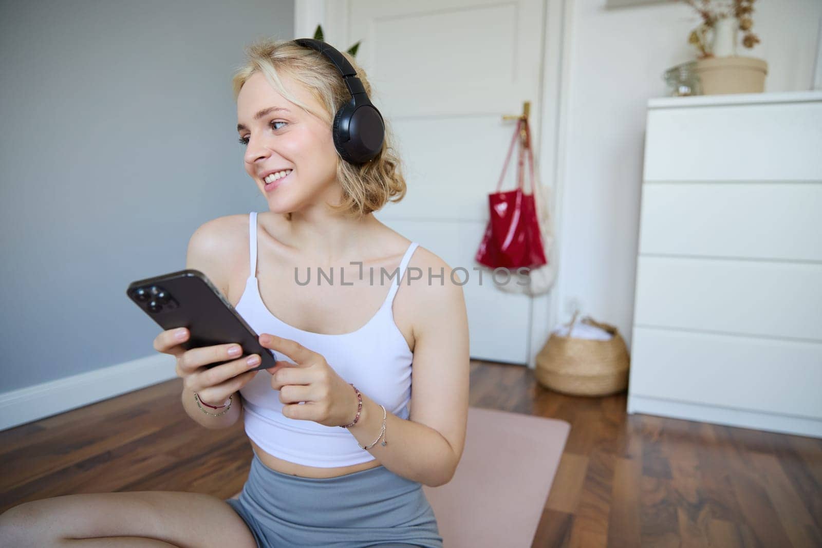 Portrait of smiling, healthy young woman in headphones, holding smartphone, working out at home, using fitness application on mobile phone by Benzoix