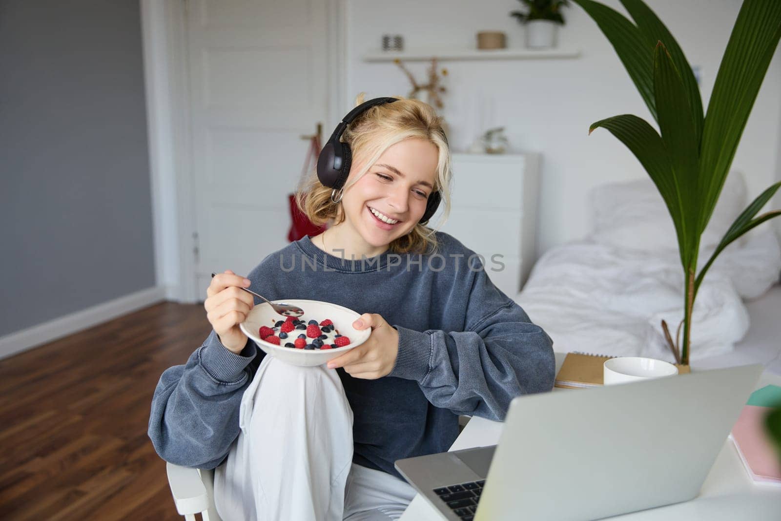 Portrait of smiling beautiful woman, sitting in room with breakfast, eating and watching tv show on laptop, laughing and looking at screen.