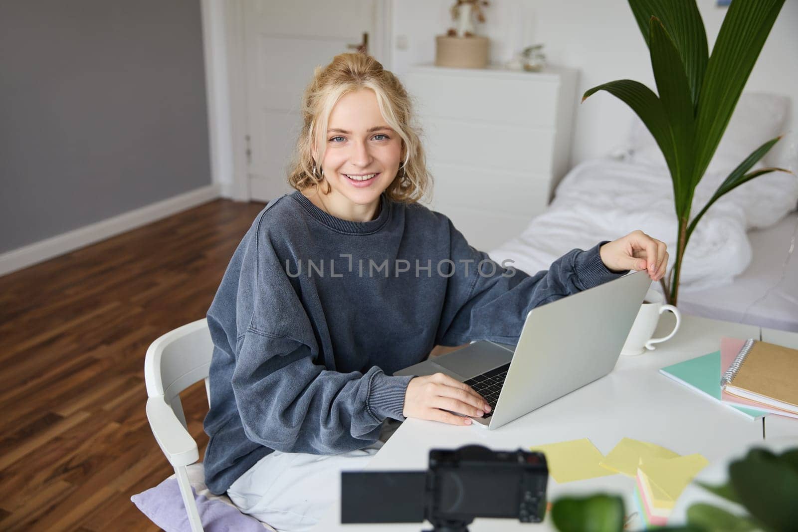 Portrait of stylish, cute young blond woman, sitting with laptop in a room, recording video on digital camera, vlogging, making lifestyle content for social media.