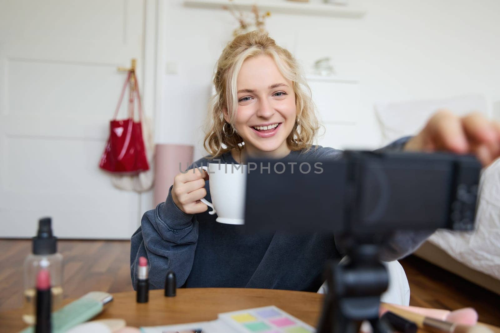 Portrait of cute smiling woman, blond girl drinks tea and records a casual, lifestyle video blog, vlogger sits in a room with camera and stabiliser, holds cup of coffee and talks.