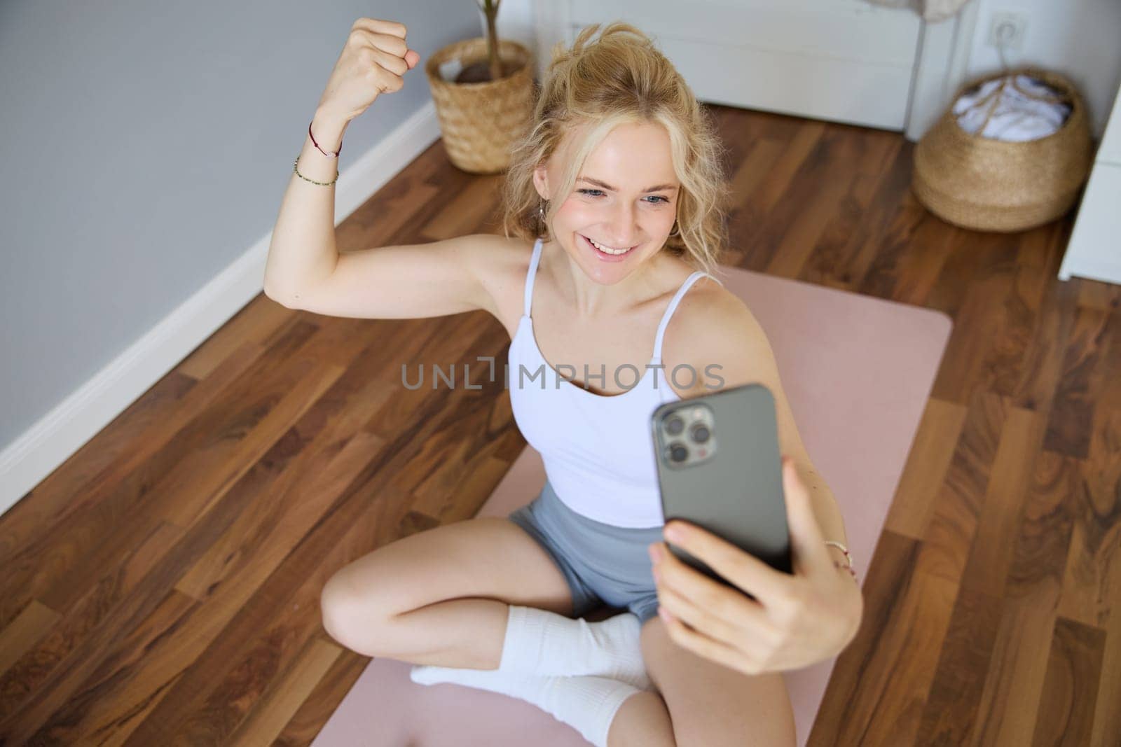 Portrait of blond young woman, fitness training, sitting on yoga mat, taking selfies during workout at home, showing muscles, flexing biceps.