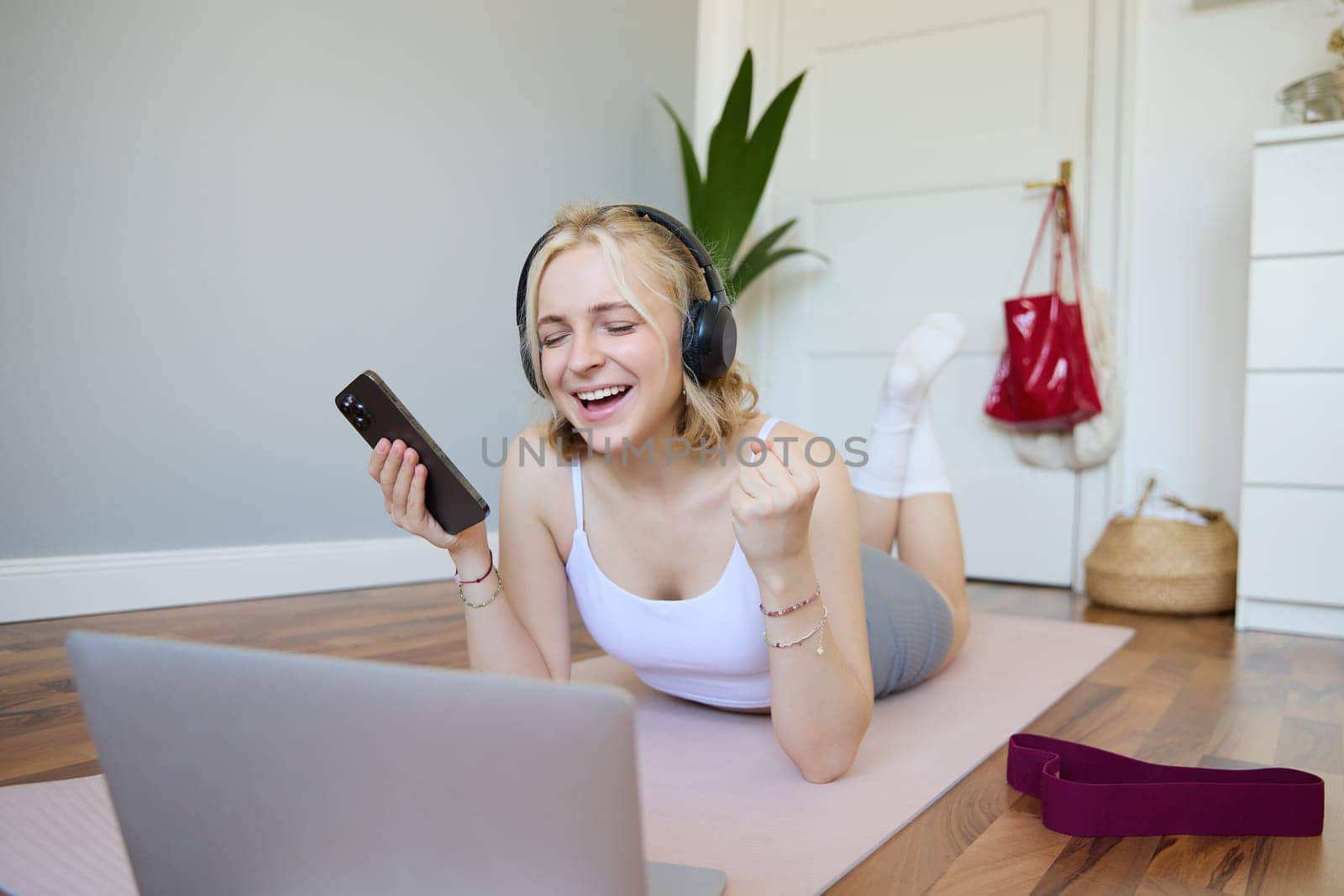 Portrait of happy blond woman in headphones, lying on rubber mat, listens to music in headphones after workout training session, using laptop by Benzoix