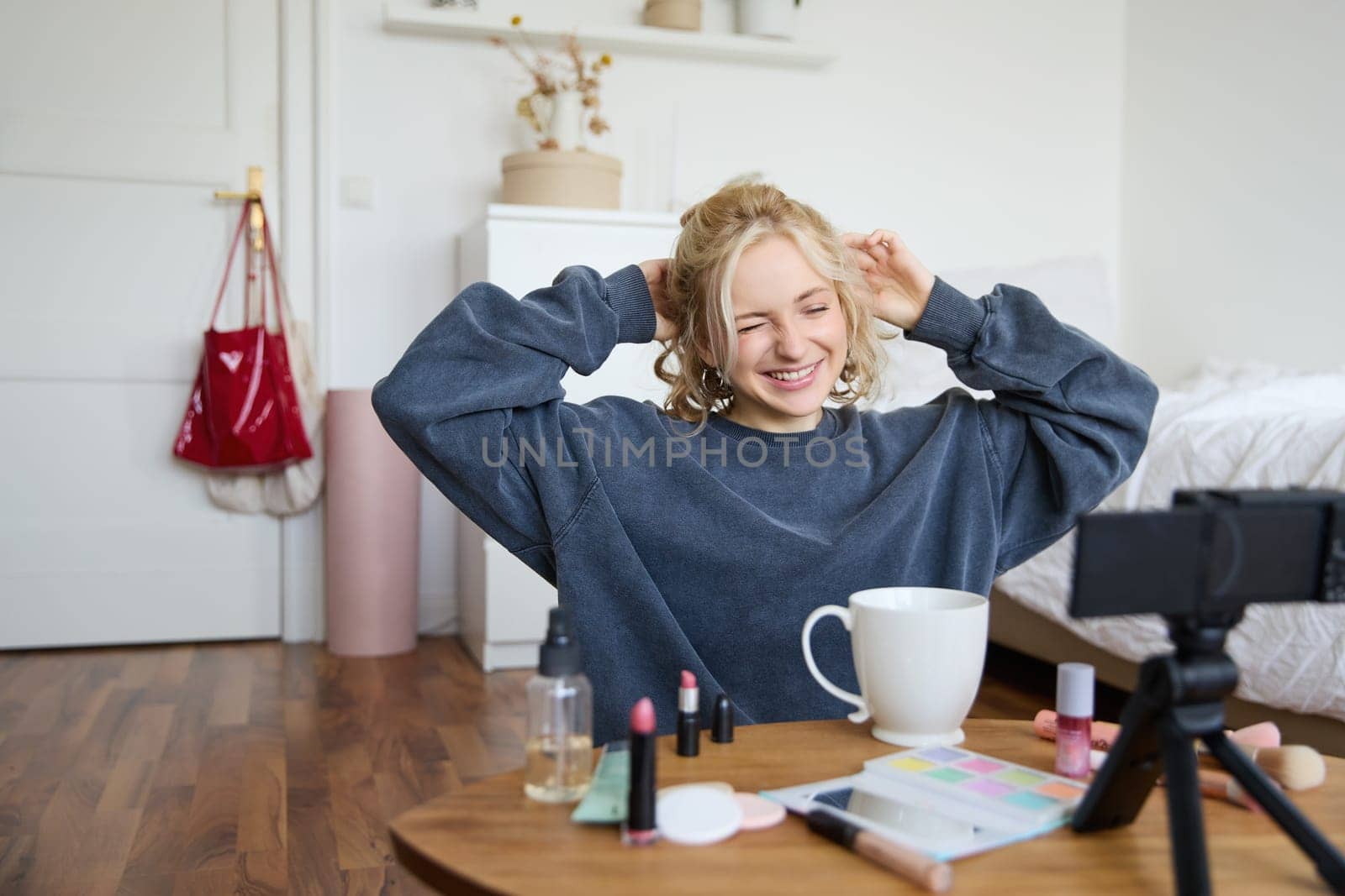 Portrait of cute smiling woman, blond girl drinks tea and records a casual, lifestyle video blog, vlogger sits in a room with camera and stabiliser, holds cup of coffee and talks.