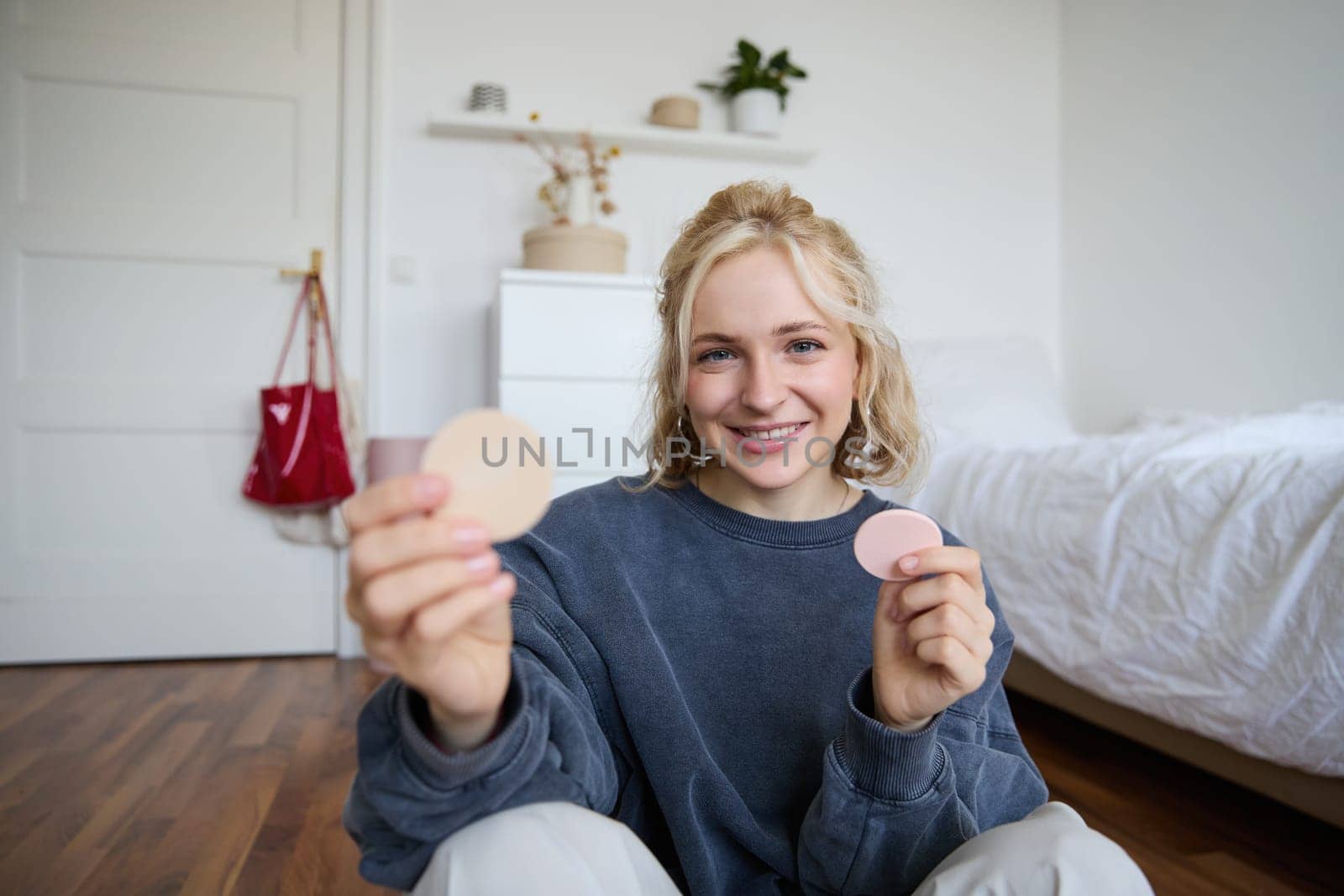 Image of smiling blond woman, vlogger, showing makeup products, beauty items, recommending to followers, sitting on floor in bedroom.