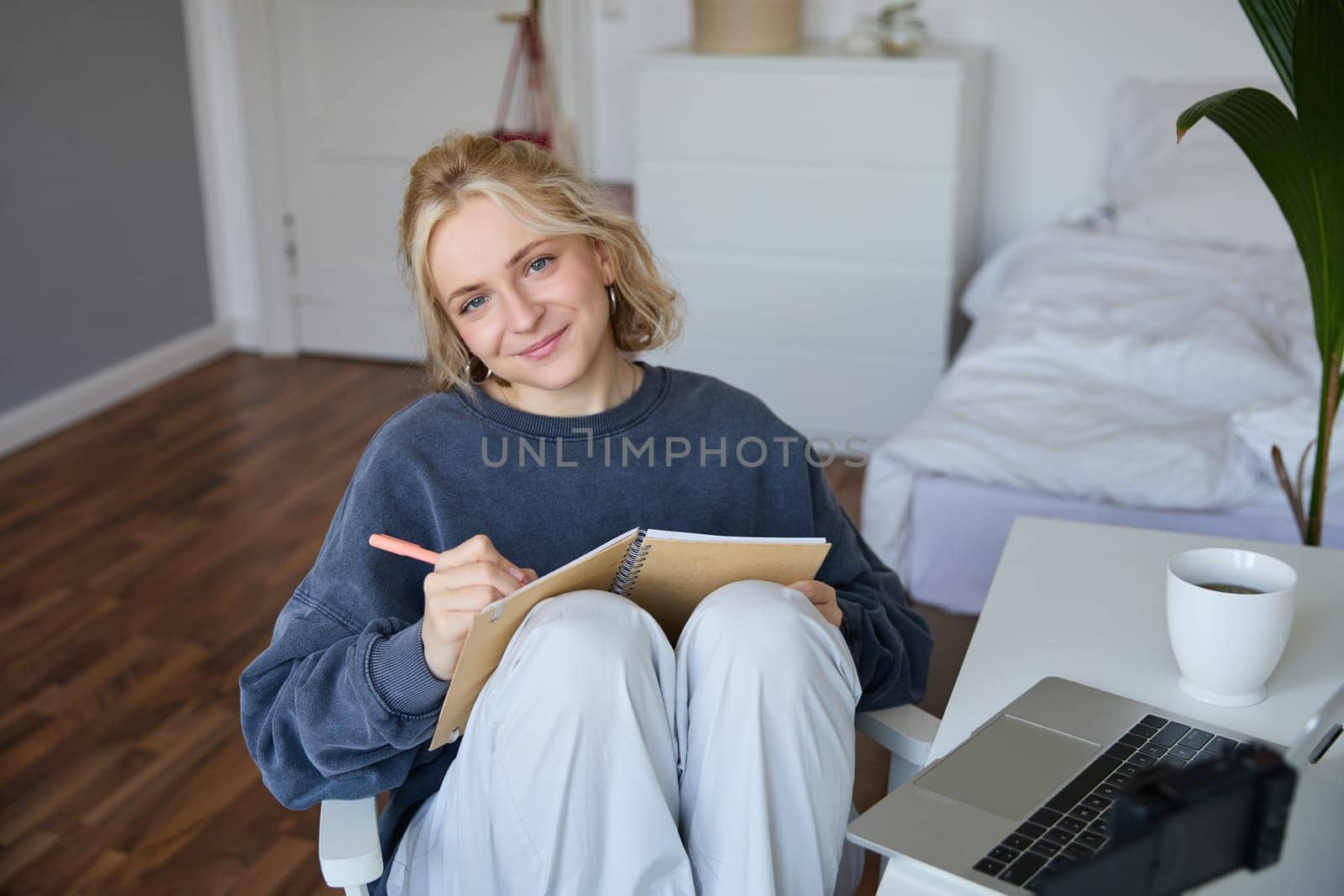 Portrait of smiling cute woman, lifestyle blogger, sits in her room with daily journal or planner, records video on digital camera, creates content for social media about daily routine.