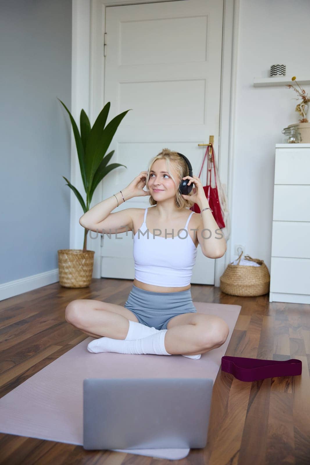 Vertical shot of smiling young woman using video tutorials for workout at home, sitting with laptop in wireless headphones on yoga mat, follows fitness instructor on social media, repeats exercises by Benzoix