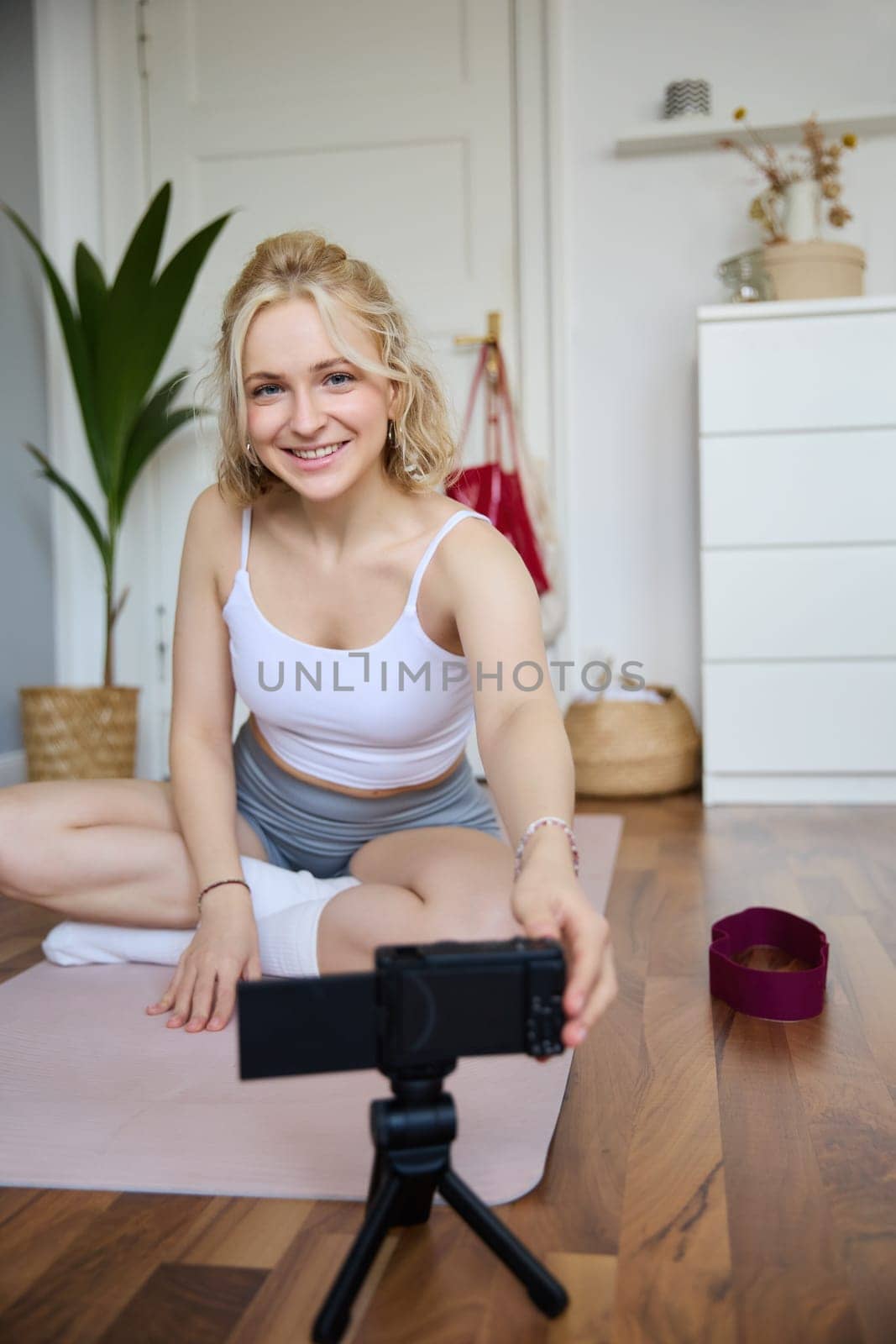 Portrait of young woman personal yoga instructor, recording workout video at home, using digital camera to vlog her exercises, using rubber mat.