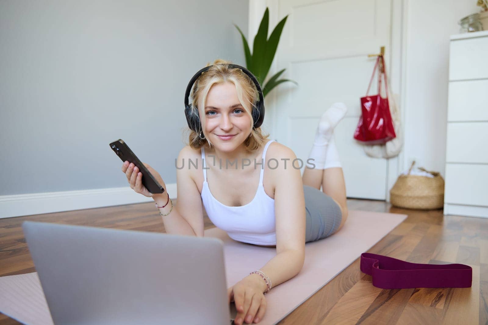 Portrait of young sporty woman, connects to online workout training session, doing exercises, lying on rubber mat, listens to fitness instructor in headphones, holding mobile phone.