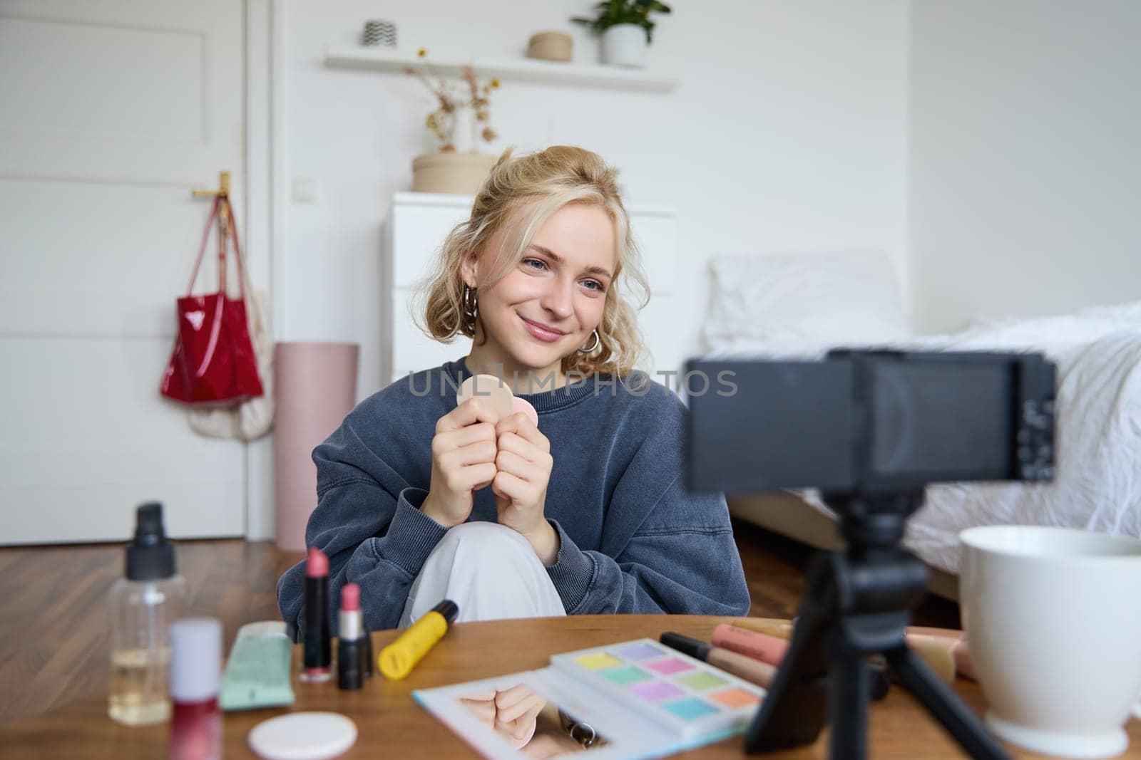 Portrait of beautiful and stylish young woman, vlogger recording video on her digital camera in a room, showing beauty products, making makeup tutorial for followers on social media.