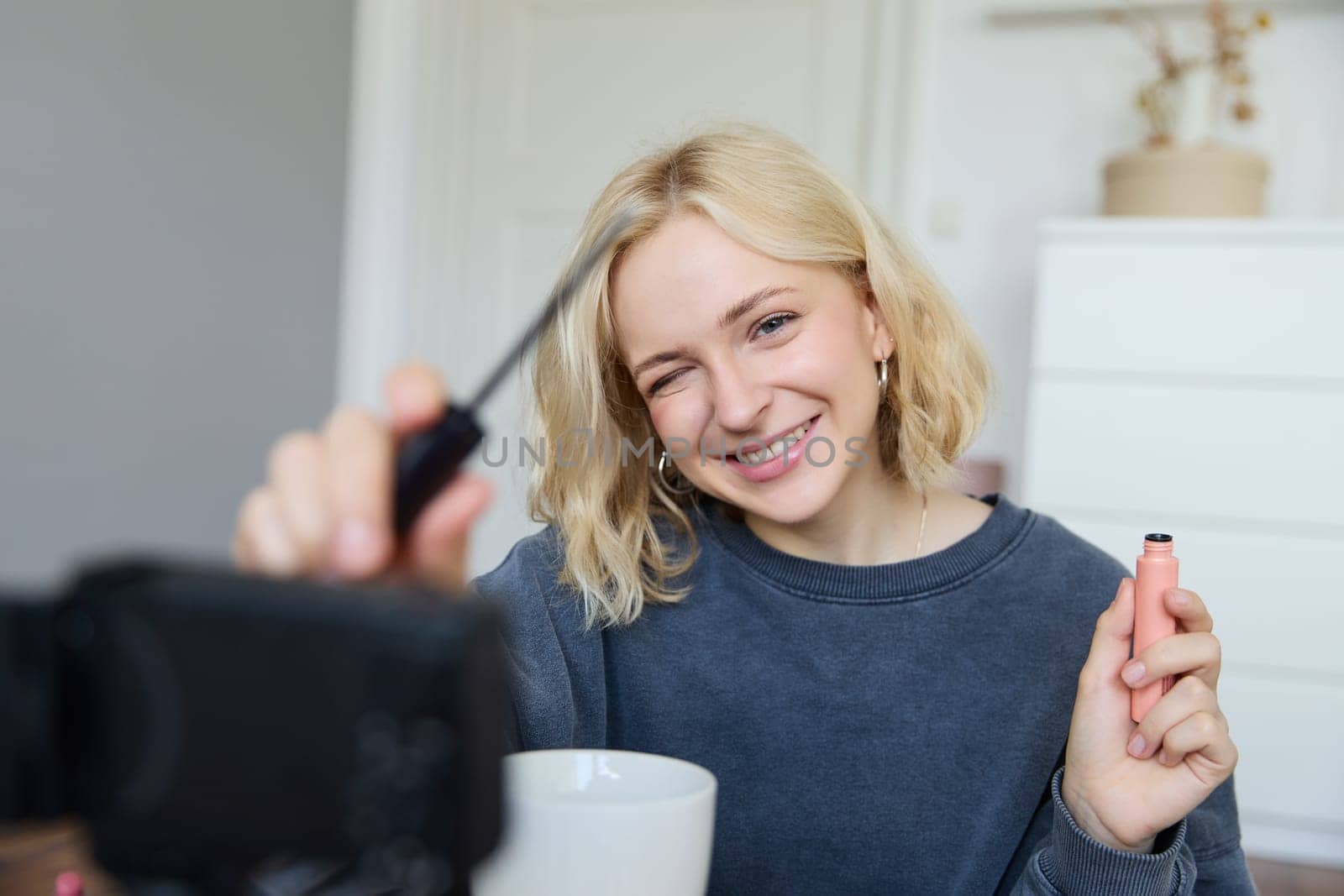 Smiling young beautiful lifestyle blogger, girl records a beauty vlog in her room, using professional video camera, showing makeup cosmetics to her followers, creates content for social media.