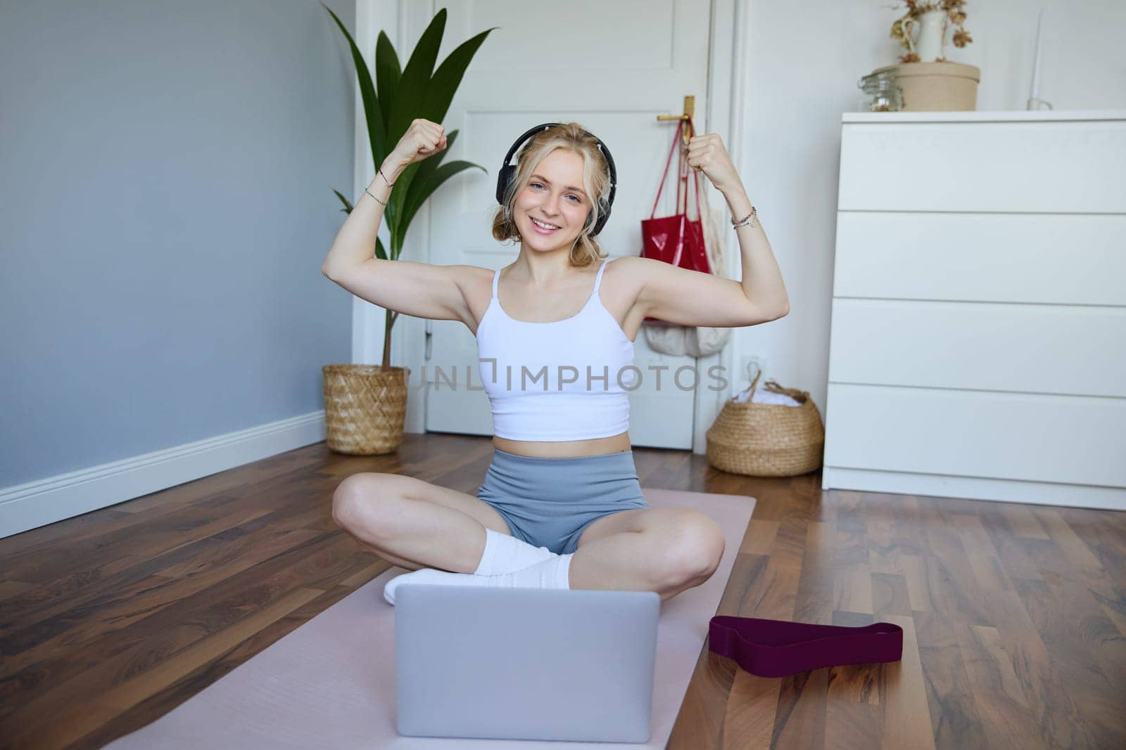 Portrait of fit and healthy, sporty woman showing her muscles on camera, wearing wireless headphones, using laptop for home workout training.