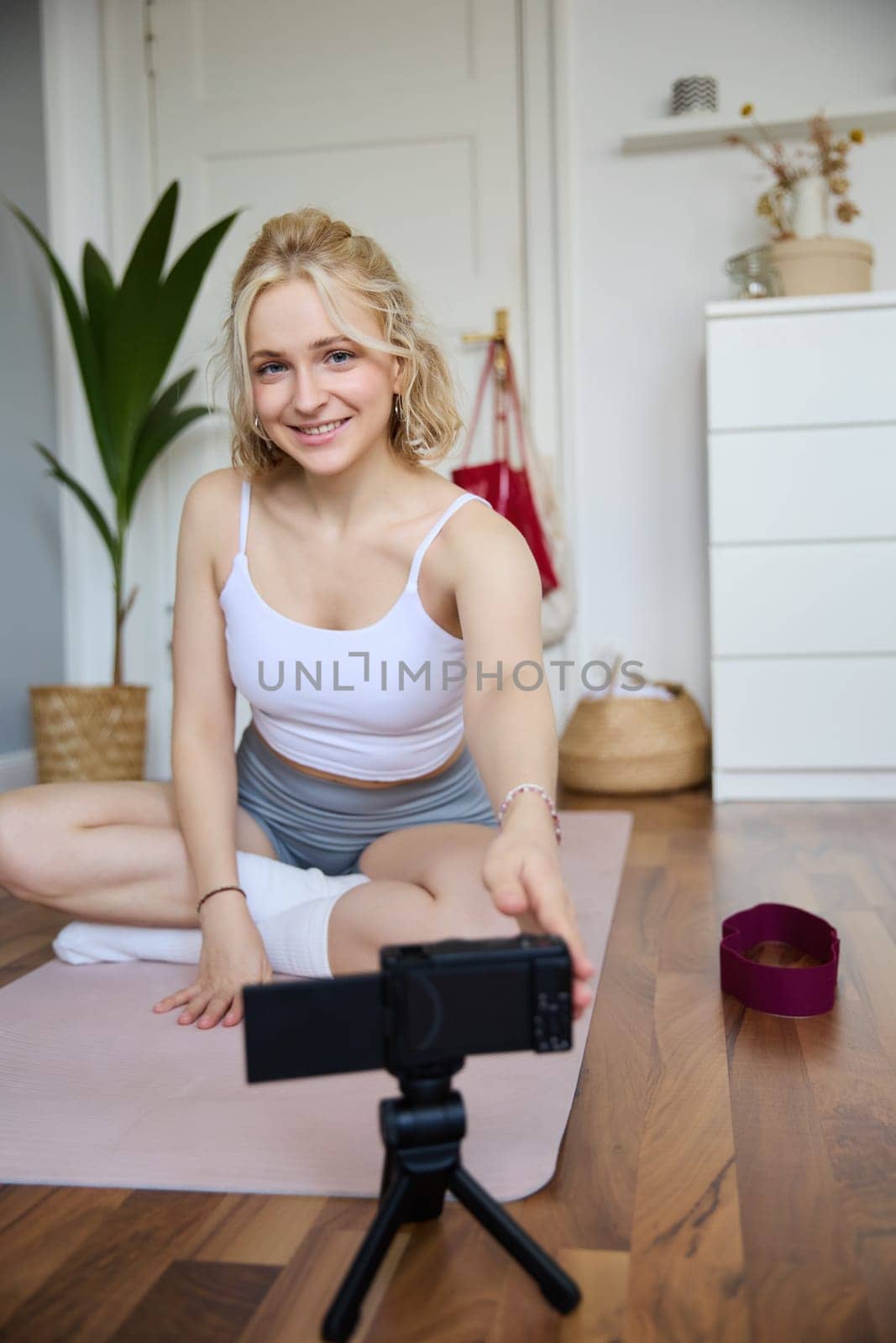 Vertical shot of woman recording her workout on digital camera, making a video about yoga and fitness at home, sitting on rubber mat in activewear.