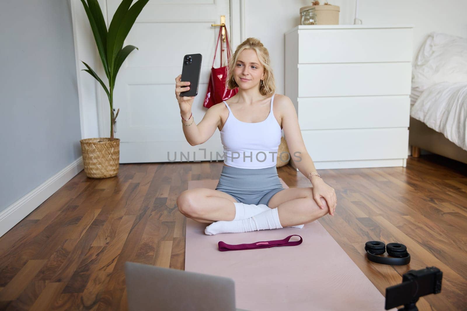 Portrait of beautiful lifestyle blogger, recording herself working out at home, sitting on rubber mat doing yoga in her room, taking selfie on smartphone, using digital video camera.