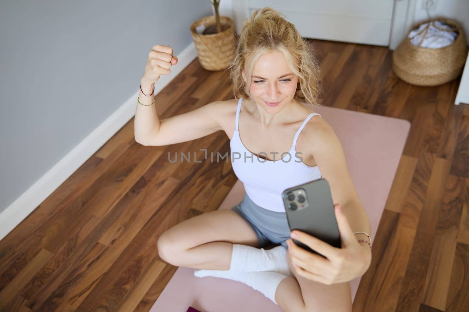 Young sporty fitness trainer, yoga instructor does live stream, takes selfies on smartphone while sitting on rubber mat at home, recording her workout training session by Benzoix