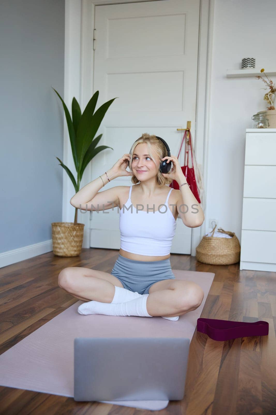 Portrait of cute young fitness woman, connects to online training session via laptop, wearing wireless headphones during workout, sits at home on rubber yoga mat.