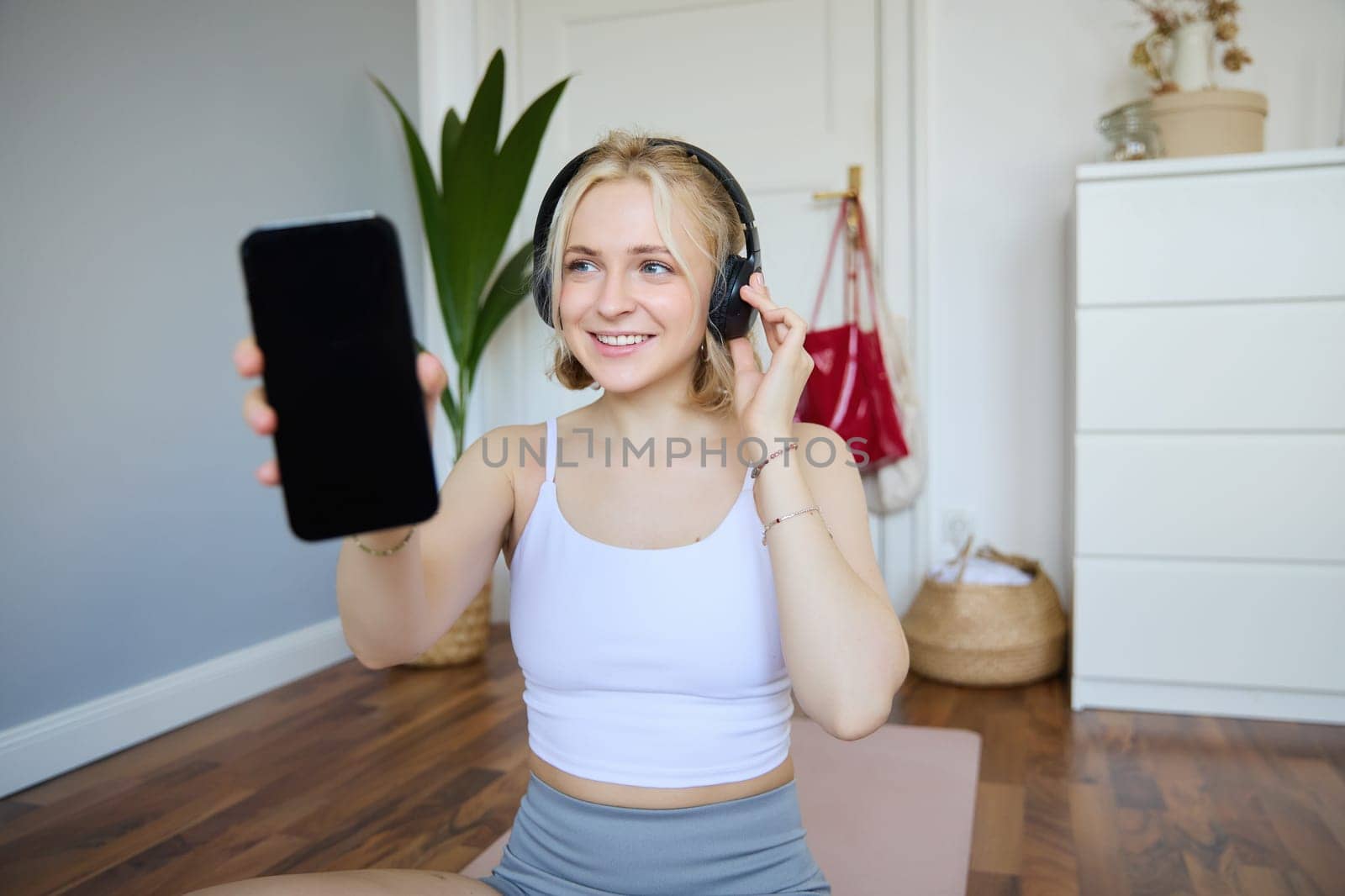 Portrait of fit and healthy woman workout at home, wearing headphones, showing smartphone screen, empty dark display by Benzoix