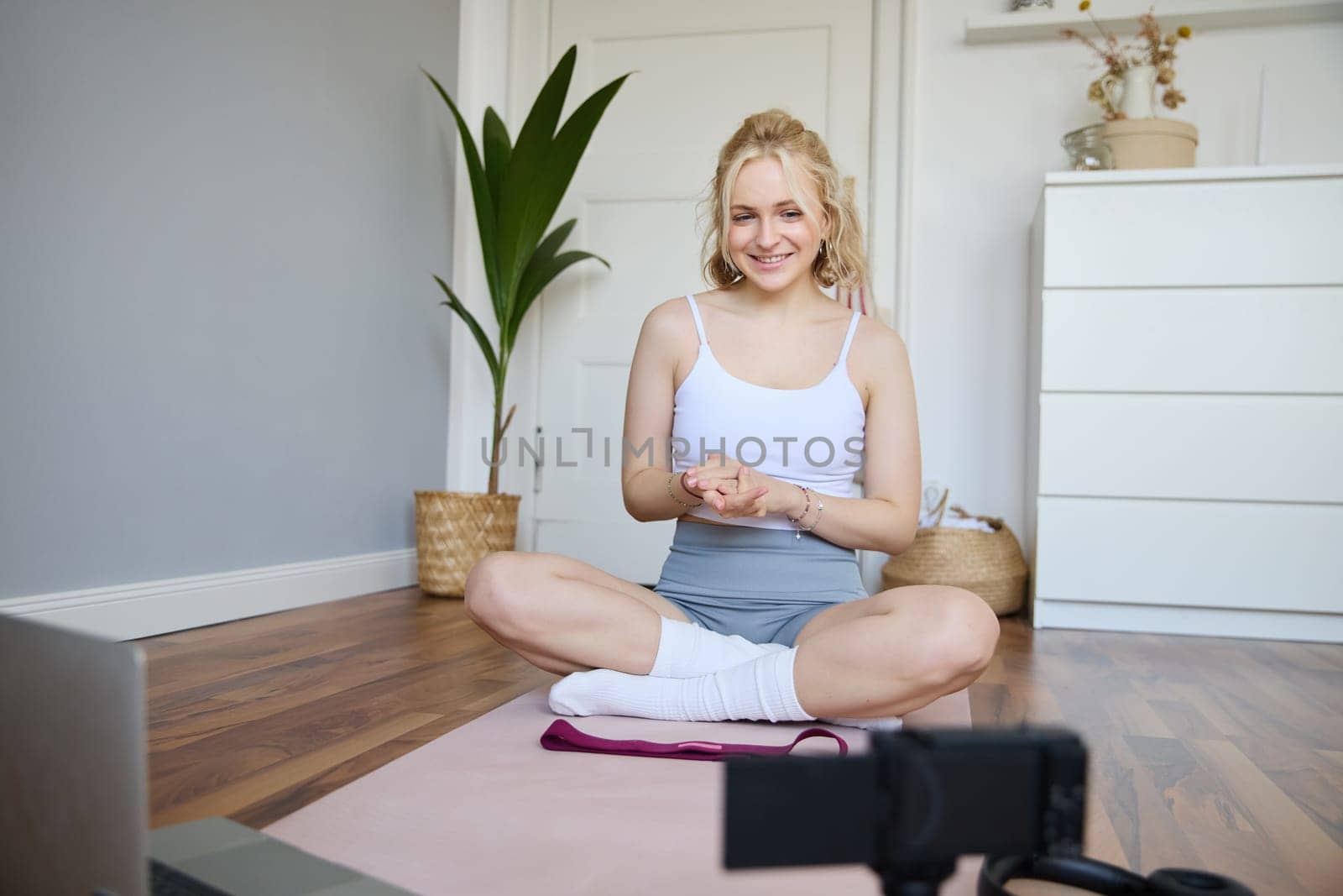 Portrait of young charismatic fitness trainer, girl blogger records video on digital camera, talks about health and workout, doing exercises on rubber mat in a room at home.