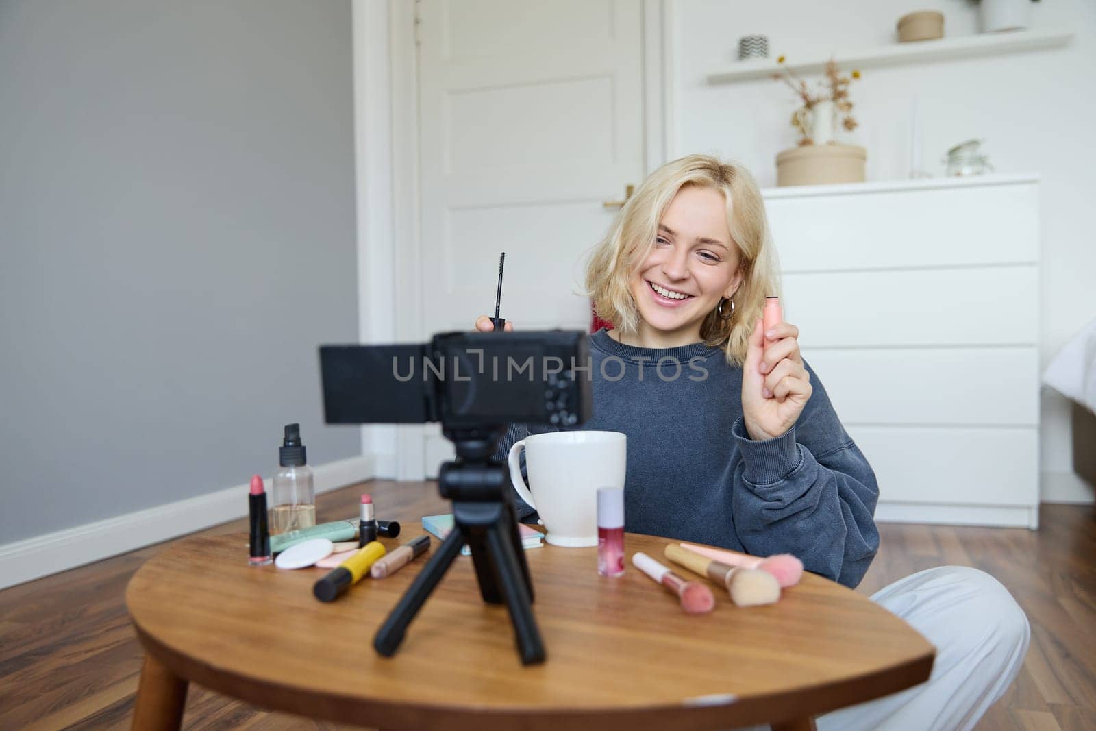 Image of cheerful, beautiful young lifestyle blogger, woman sitting on floor and recording video about makeup, holding mascara, making lifestyle content for her social media account and followers.