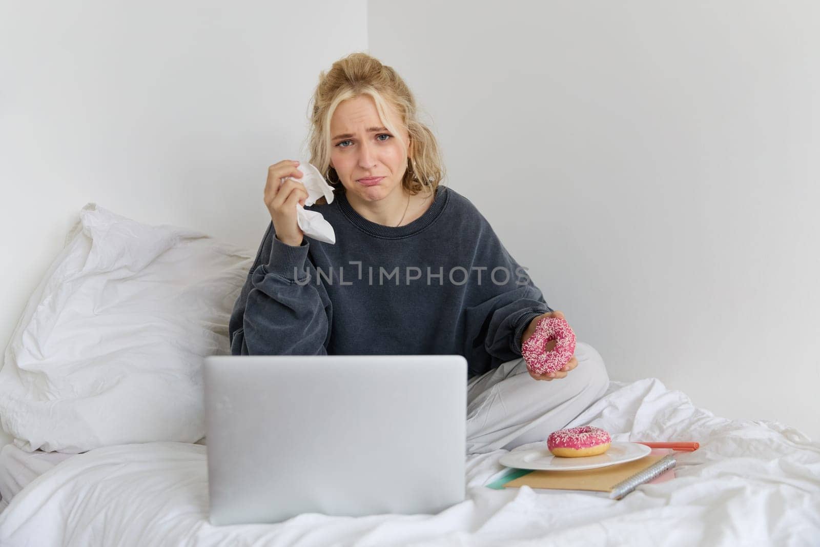 Portrait of woman watching sad movie on laptop, crying and wiping tears off, eating doughnut and drinking tea.