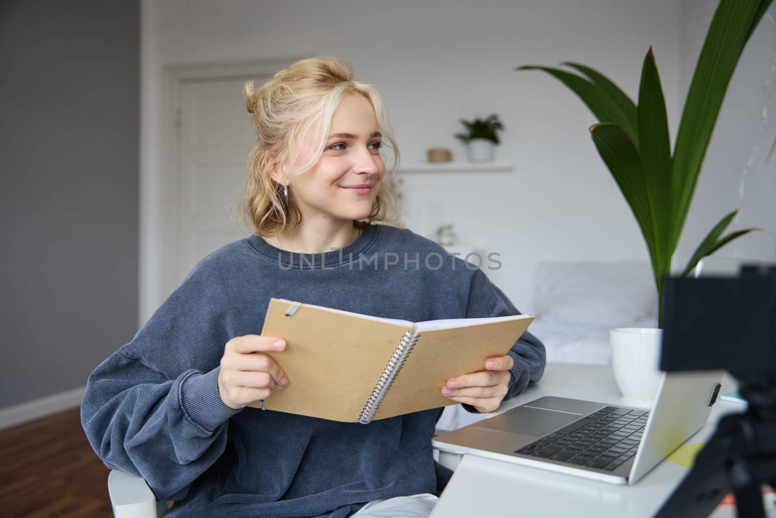 Portrait of cute young woman, teenage girl records video on digital camera, uses laptop to create lifestyle content, shows notebook, reads notes, using computer in her room.