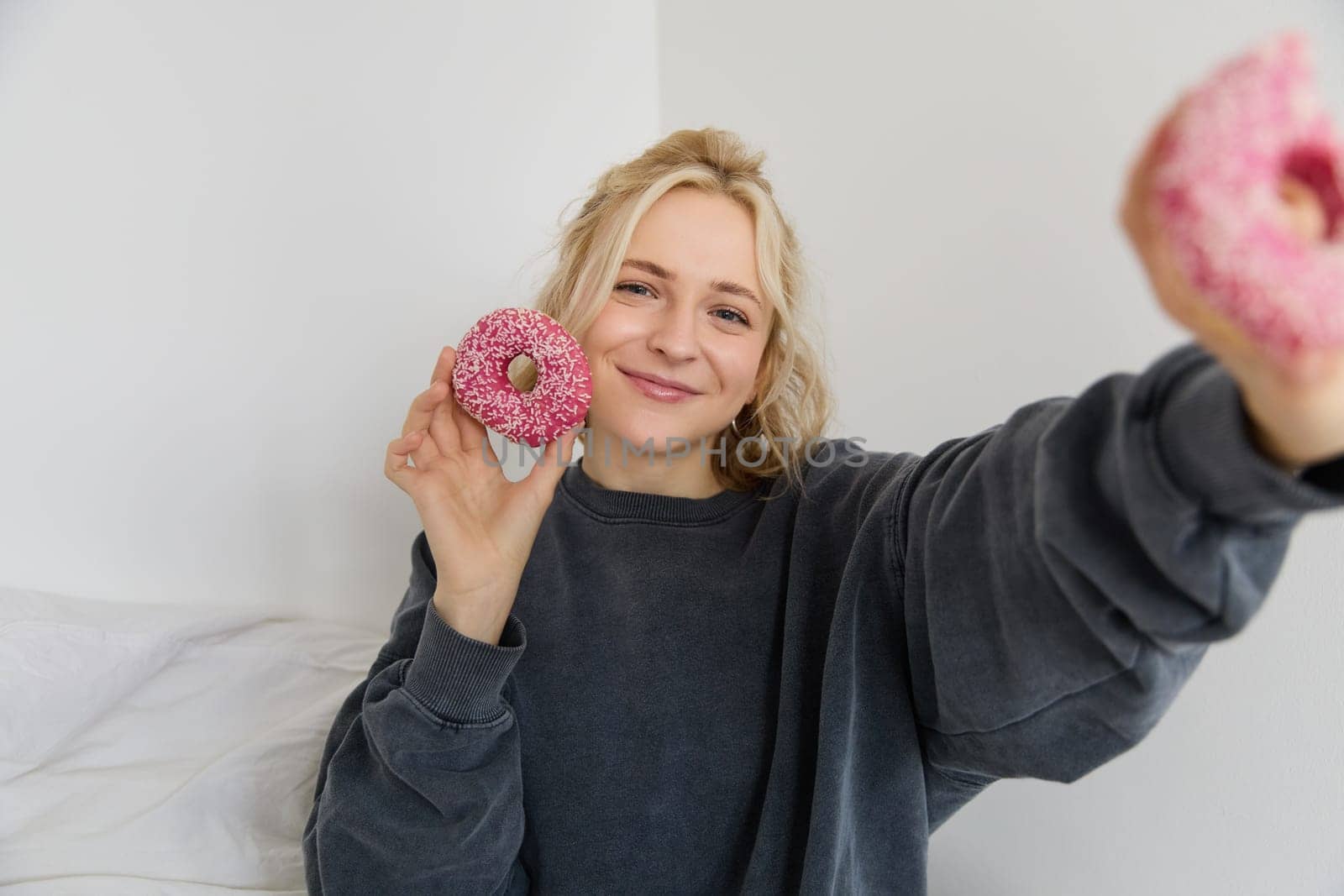 Portrait of beautiful smiling blond woman, showing two pink doughnuts at camera, eating delicious food.