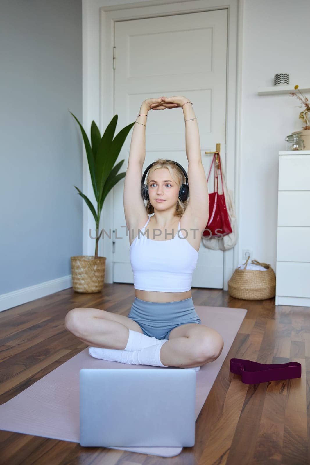 Portrait of young fitness woman doing warm-up exercises, looking at workout video on laptop, stretching arms, wearing headphones by Benzoix