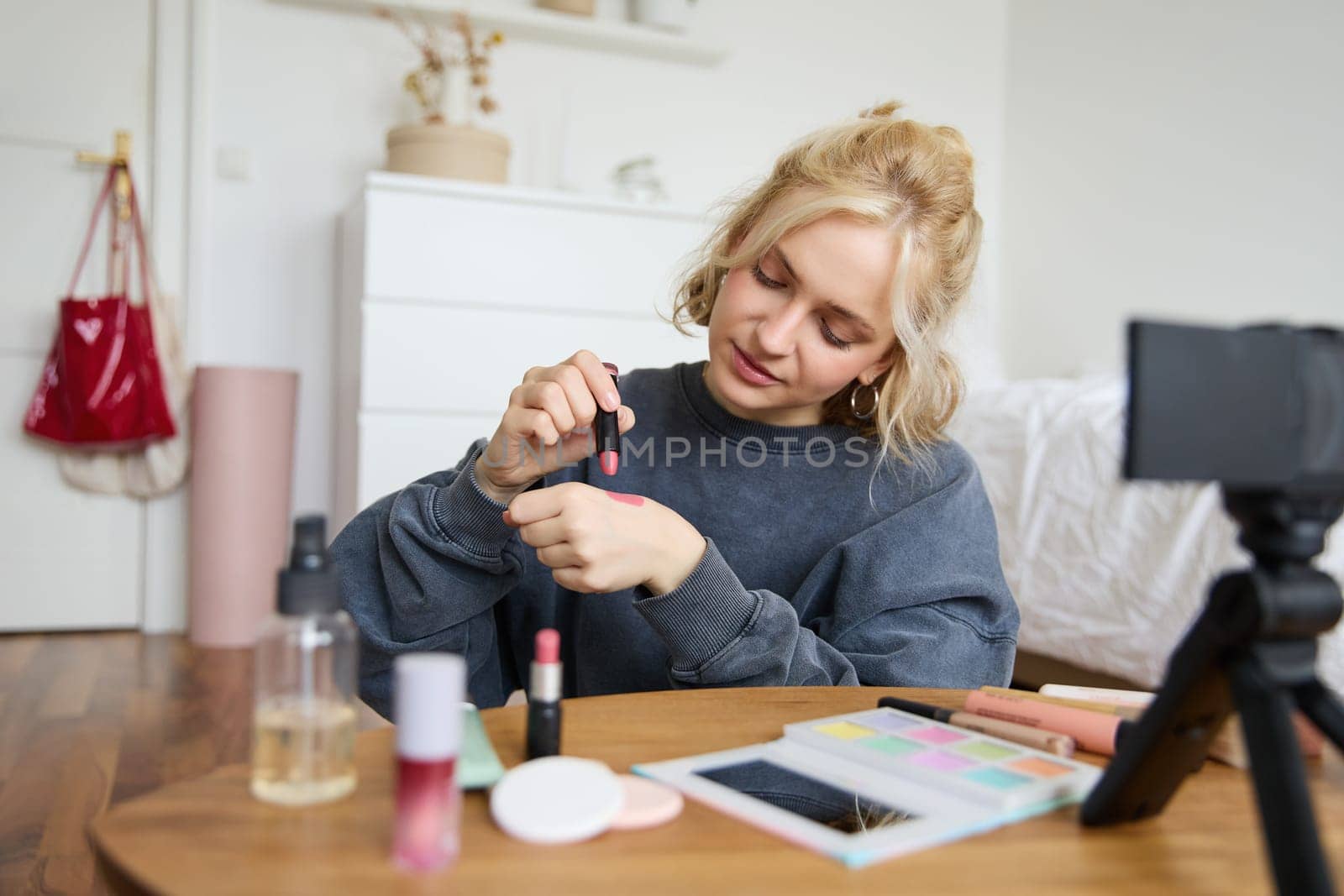 Woman beauty blogger, records video of herself sitting in a room and rating makeup products, puts on make up, holds lipstick and cosmetic brush in hand, using professional camera for content creation.