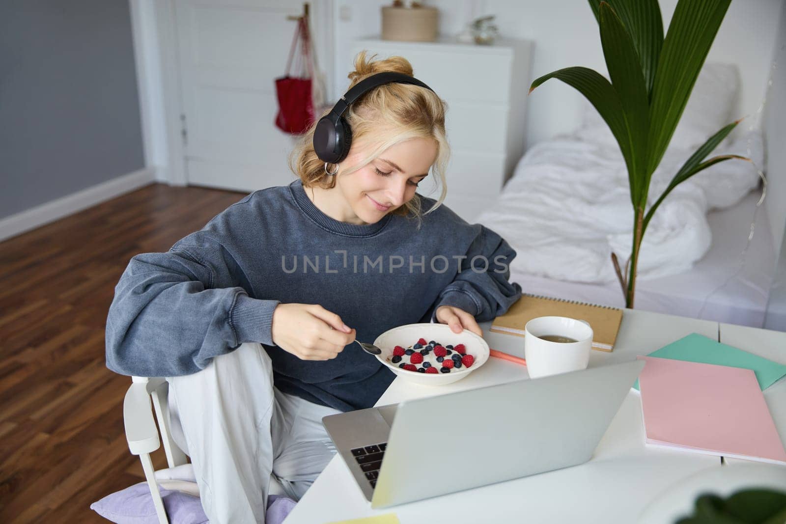 Portrait of smiling young woman, watching tv show in headphones, eating breakfast and looking at laptop screen.